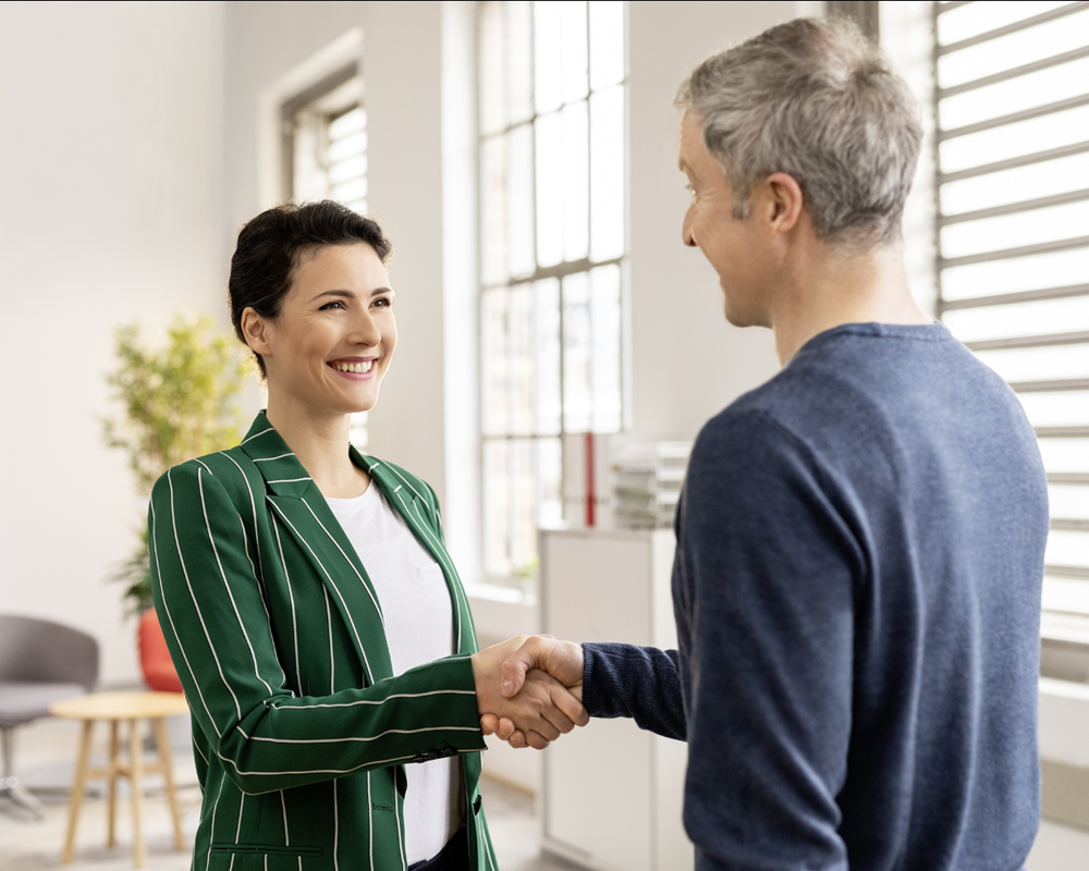 Women with short dark hair in green jacket and men with grey hair and blue pullover shaking hands in an office