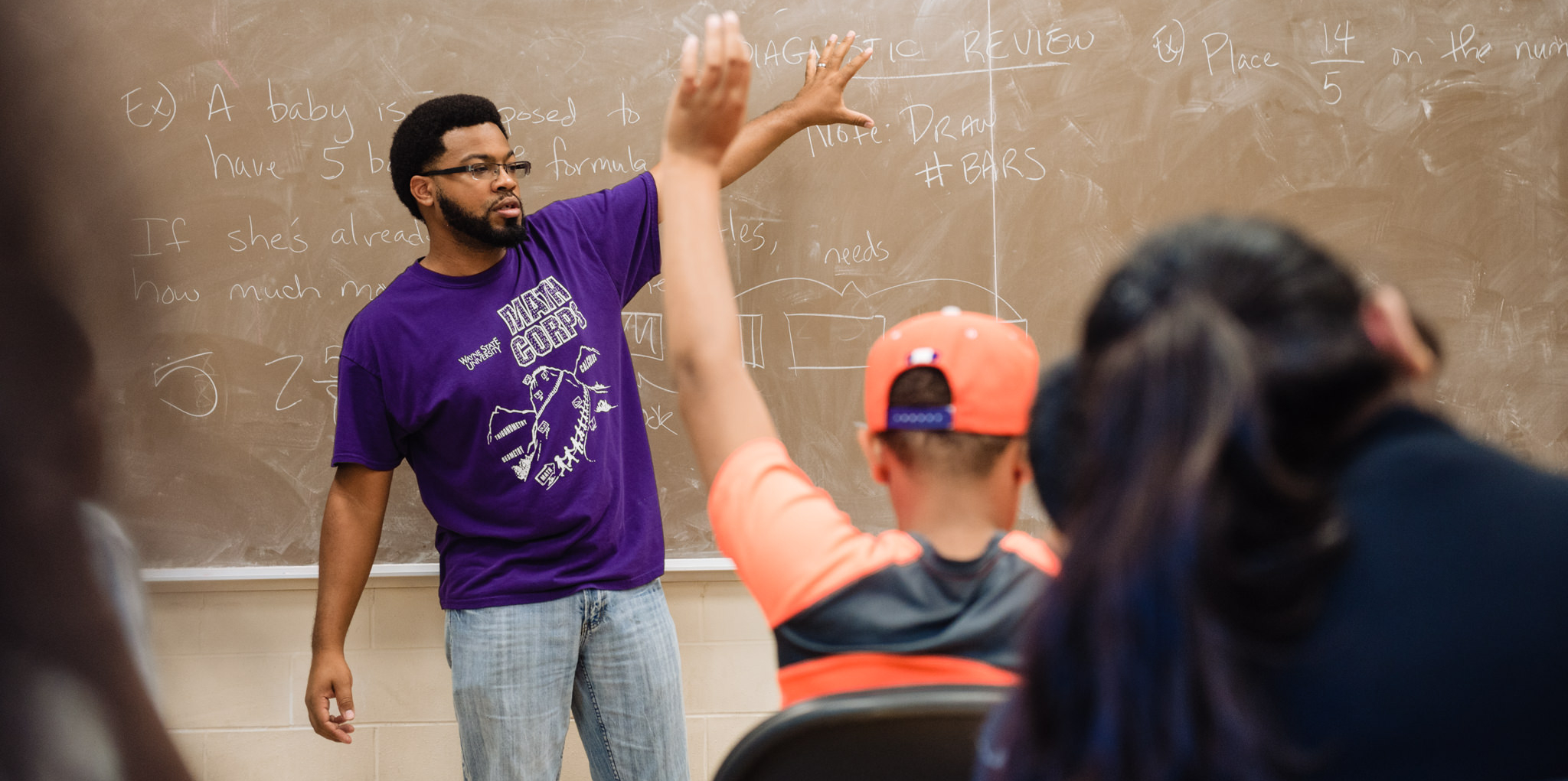 Man standing at chalkboard in classroom while student raises hand
