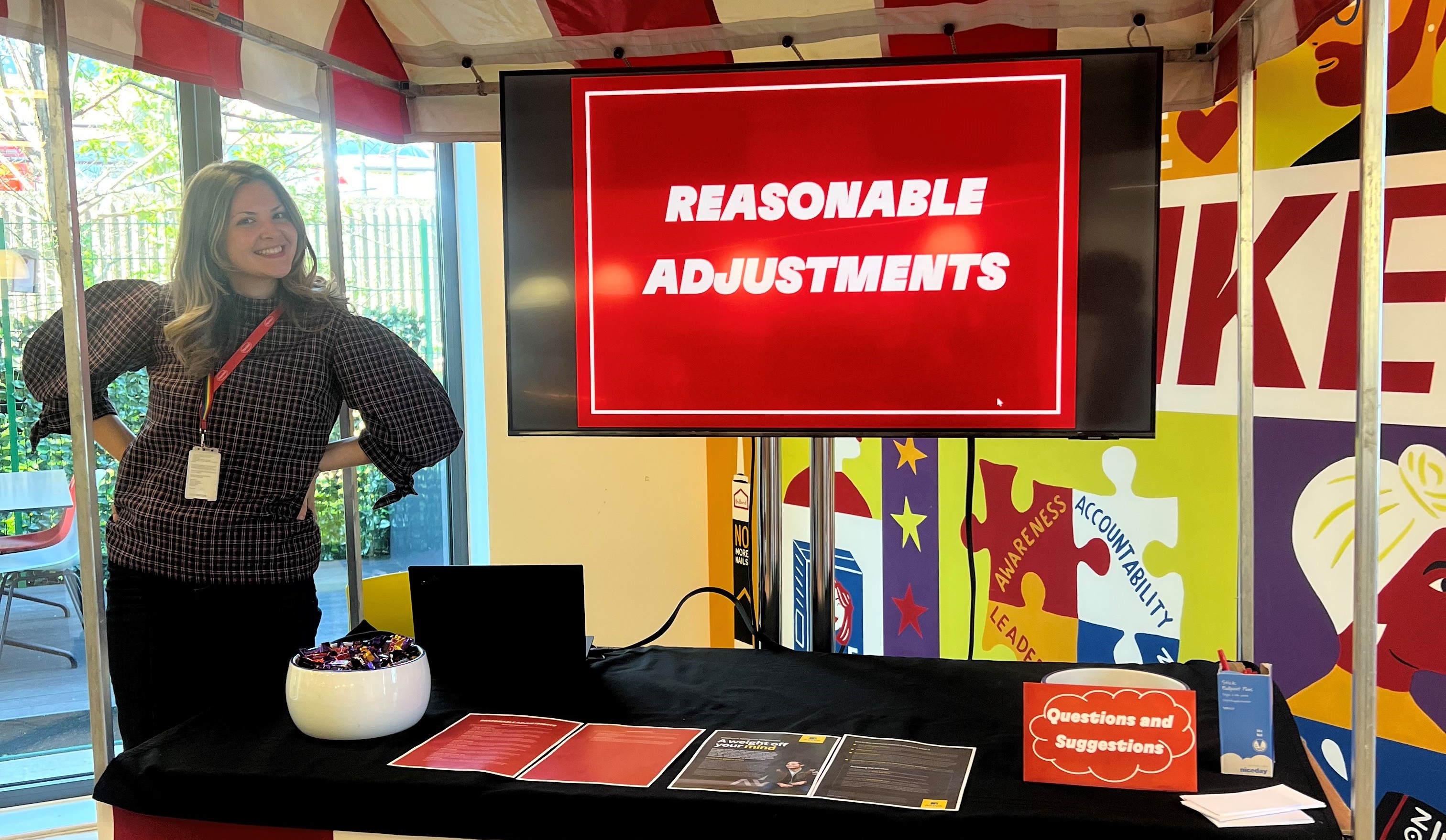 A woman poses next to a ‘Reasonable Adjustments’ slide, a sweets bowl, and a bowl with a ‘Questions and Suggestions’ sign.