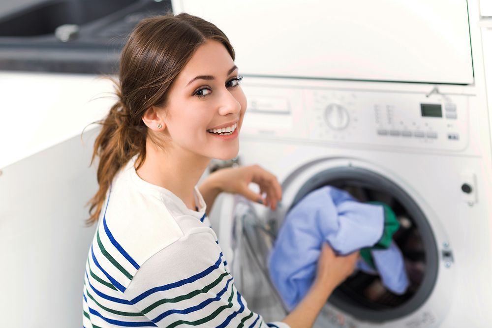 Women with long brown hair and a white pullover with blue stripes puts laundry into the washing machine, smiling at the camera.