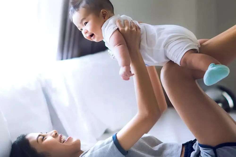 Mother lifting and playing with a smiling baby indoors in a bright home environment.