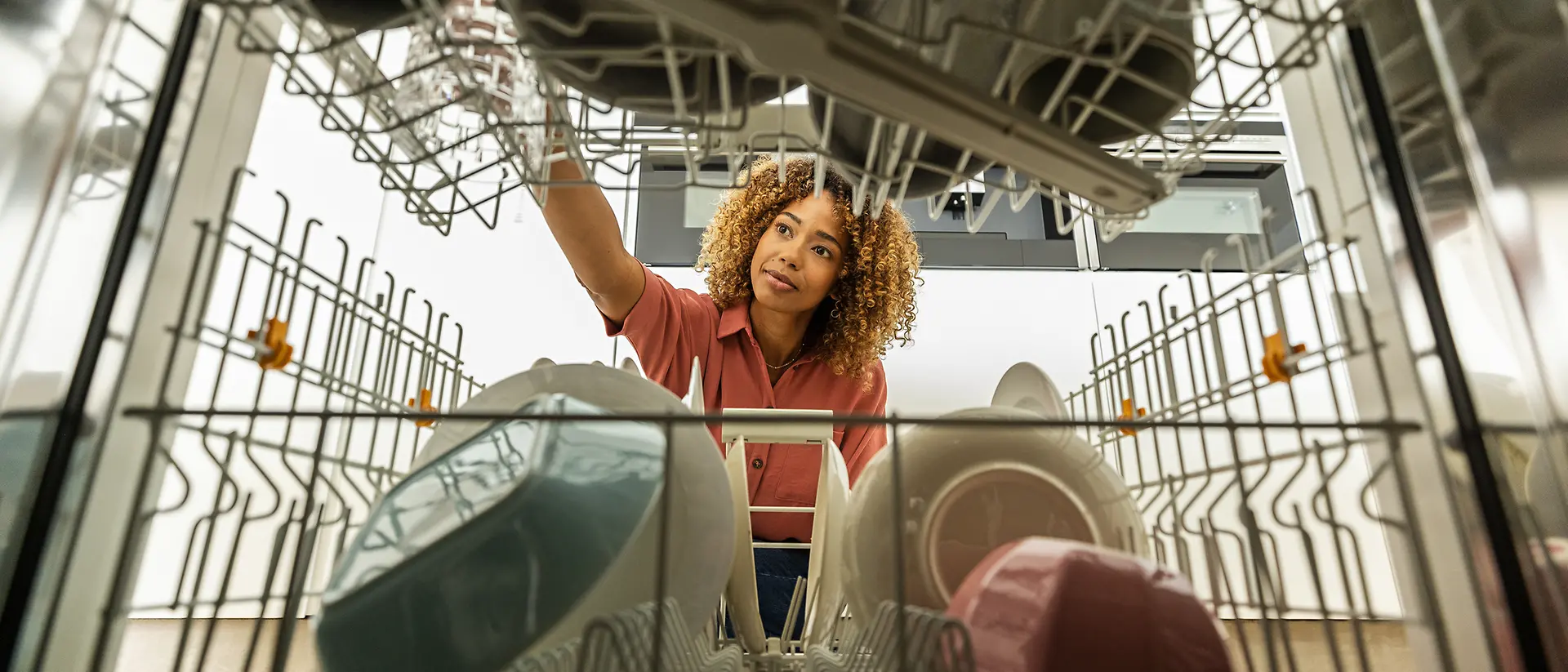 From the perspective inside a dishwasher, a young Black woman with curly hair is seen placing a glass onto the top rack, her expression is focused, and the kitchen is softly lit in the background.