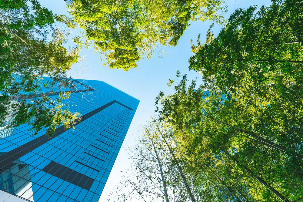 View from below a modern skyscraper that stretches into the sky and is surrounded by green trees.