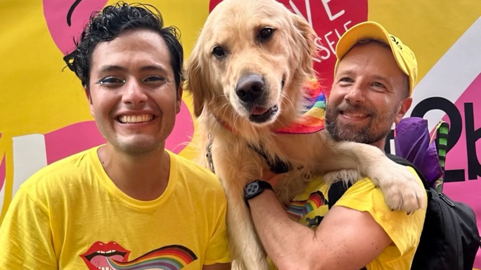 A photo of Luis and James, holding a dog, wearing got2b t-shirts and smiling into the camera at a Pride event. 