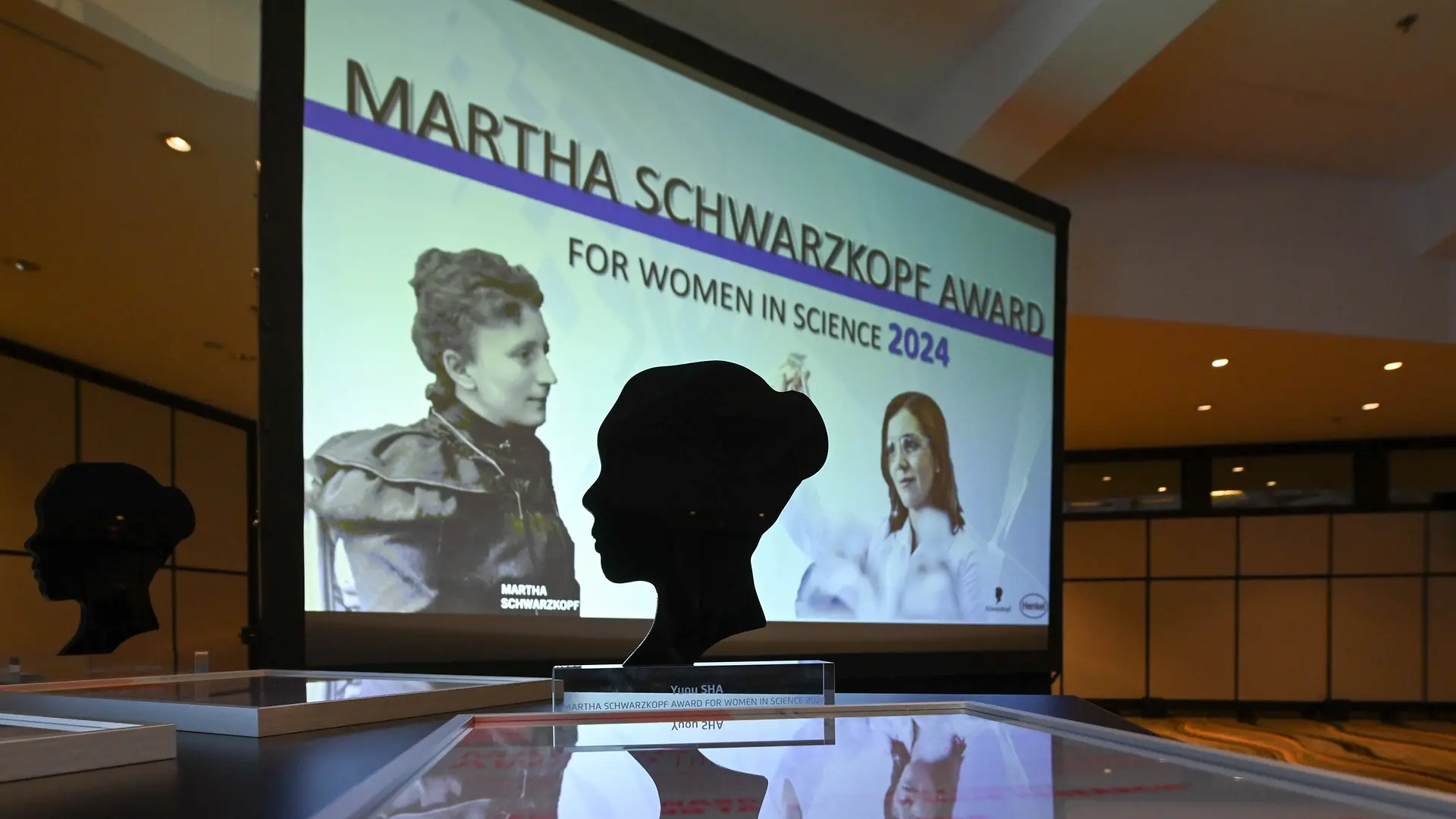 The image shows the Martha Schwarzkopf Award placed on a table. In the background, a large screen displays the text “Martha Schwarzkopf Award for Women in Science 2024.” The scene reflects the formal setting of an award ceremony.