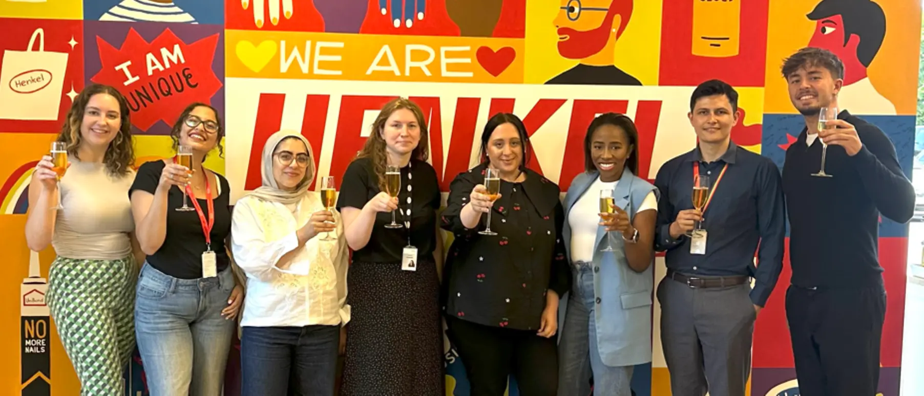 9 people raising a glass, standing in front of a colourful ‘We are Henkel’ wall.