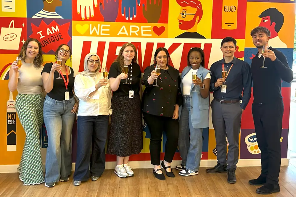 9 people raising a glass, standing in front of a colourful ‘We are Henkel’ wall.