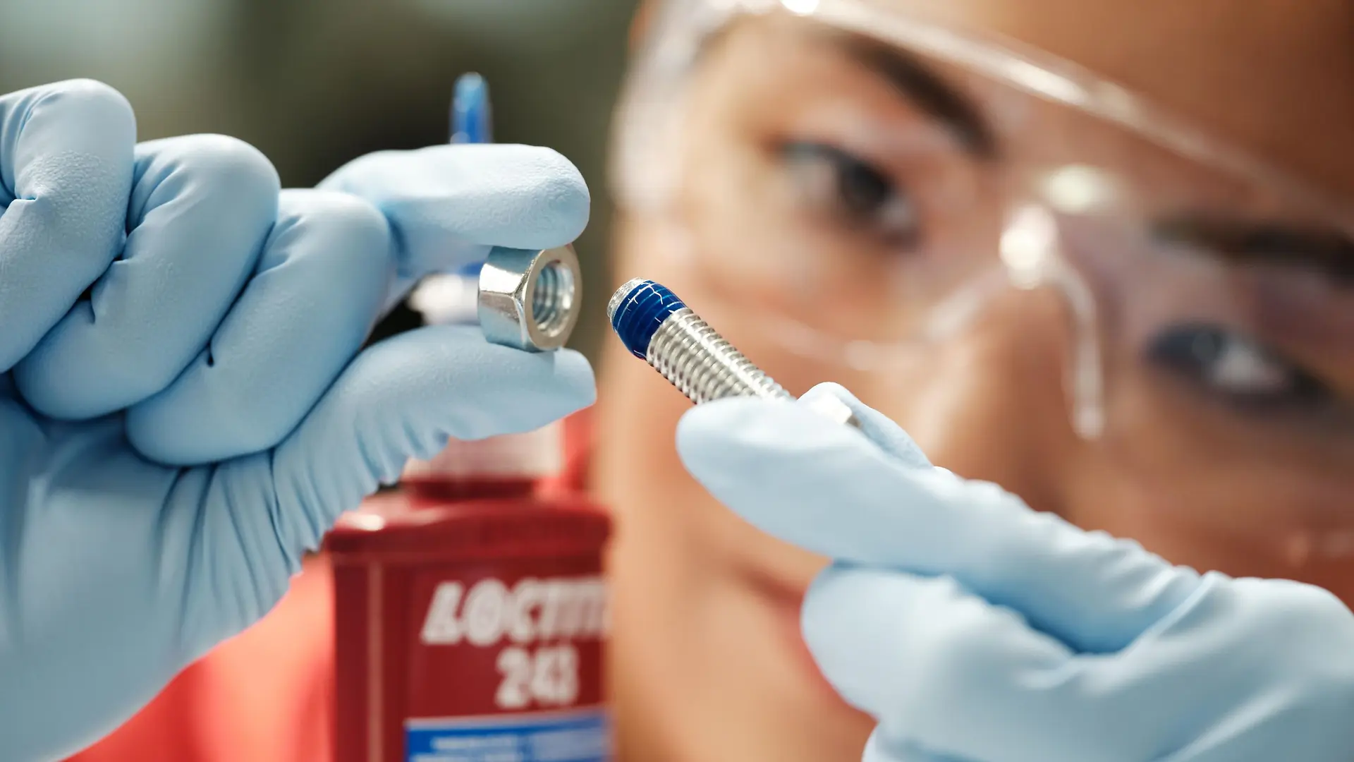 A woman's face in the background. In the front her hands with blue gloves holding a screw with blue Threadlocker.