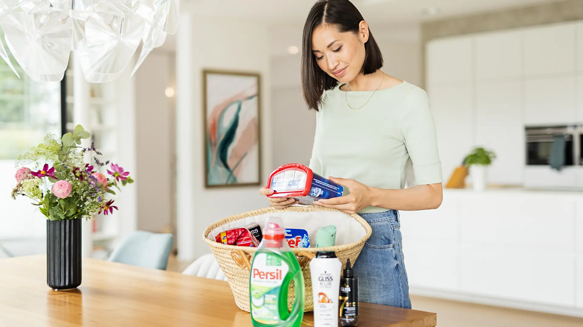 Women with a basket standing on a table that is full of Henkel products such as: Persil, Gliss, Bref, Somat, taft and more.