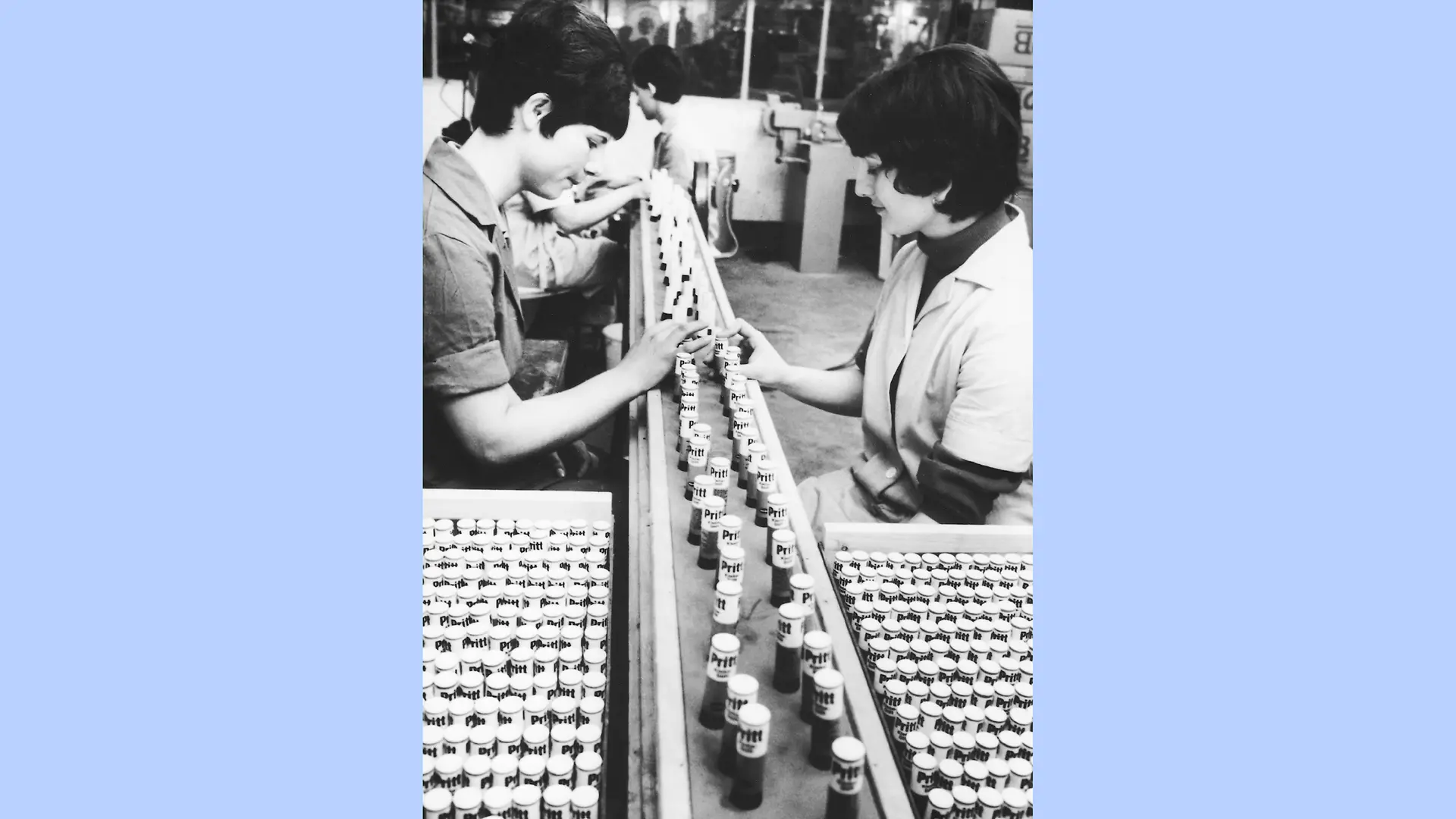 photo en noir et blanc de l’usine de conditionnement des produits Pritt. Deux femmes sont assises face à face de part et d’autre d’un tapis roulant sur lequel des sticks de colle Pritt sont disposés en rangées. À côté de chaque femme se trouve une boîte remplie de sticks Pritt supplémentaires.