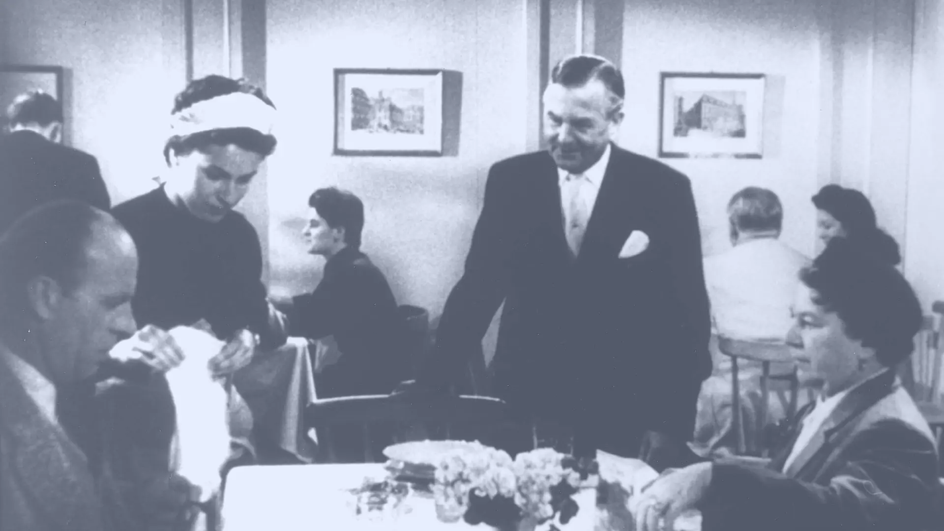 À gauche, un homme et à droite, une femme assise à une table de restaurant. À côté de l’homme se tient une domestique portant une coiffe blanche. À côté de la femme se trouve un serveur.