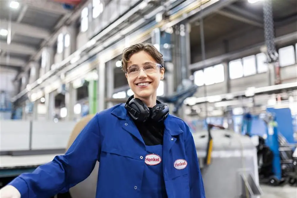 Smiling Henkel employee wearing a blue work coat and safety headphones, standing in an industrial production hall, representing expertise and innovation in manufacturing.