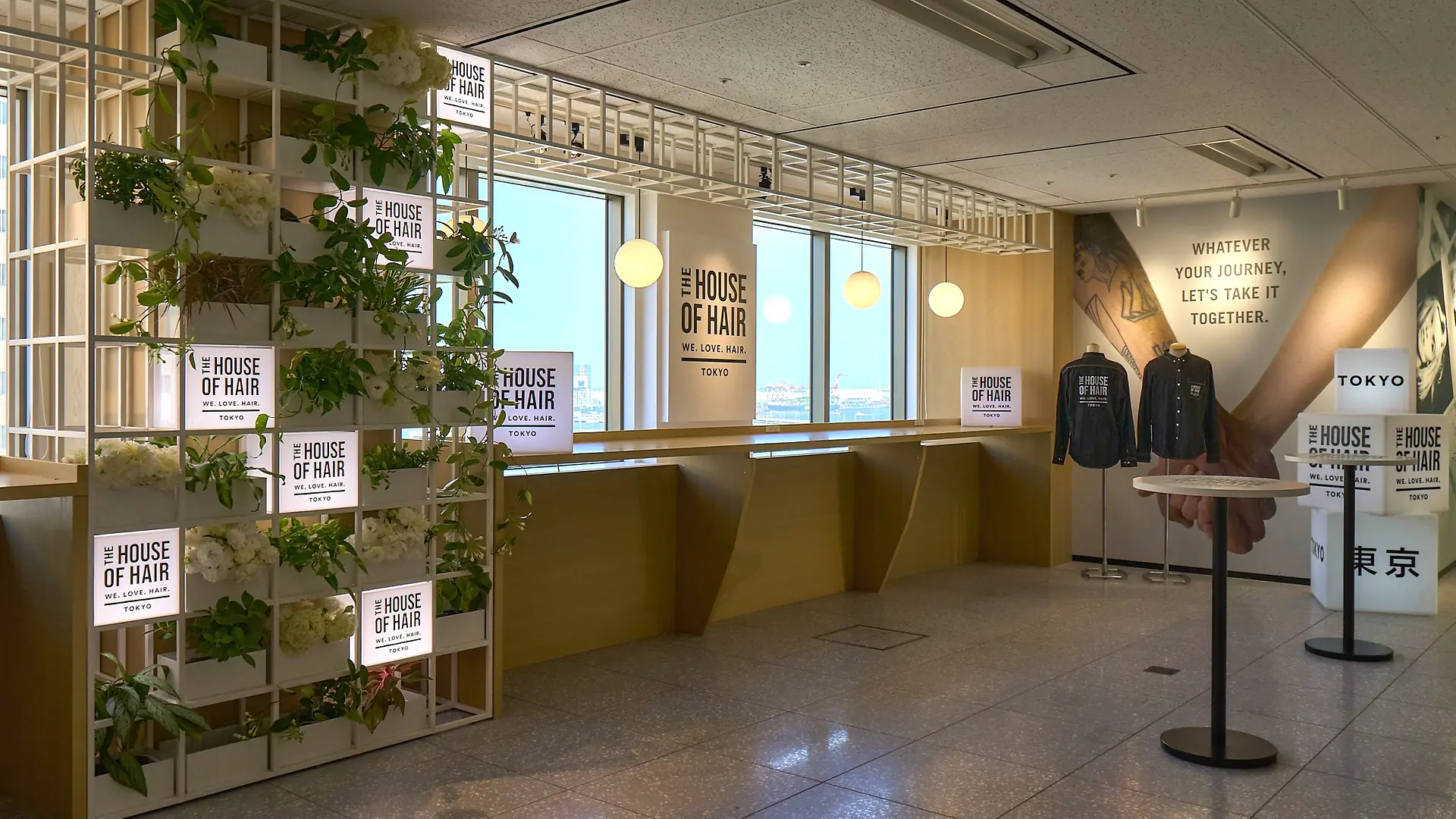 Un espace intérieur moderne avec de grandes baies vitrées laissant entrer la lumière naturelle, comprenant un comptoir en bois le long des fenêtres et des étagères décoratives avec des plantes vertes et des enseignes lumineuses indiquant « House of Hair Tokyo ». Sur la droite, un mur affiche une citation motivante : « Quel que soit le voyage, faisons-le ensemble », ainsi que deux sweats à capuche noirs présentés sur des supports. L’ensemble présente un design épuré et contemporain, avec des tons neutres et des touches de verdure.