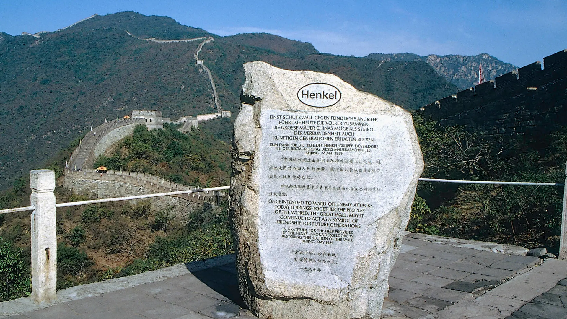 Une pierre commémorative célébrant le soutien de Henkel à la restauration d’une section de la Grande Muraille de Chine. À l’arrière-plan, la Grande Muraille serpente à travers une zone montagneuse boisée.