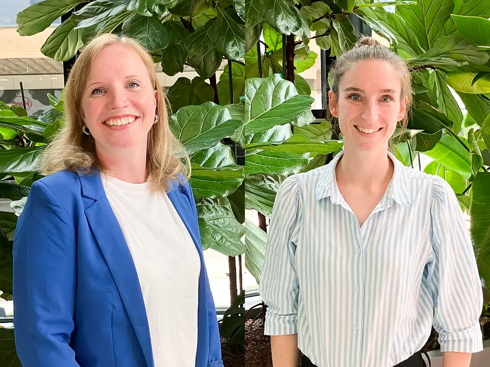 Two recruitment partners standing indoors in front of large green plants. One is wearing a blue blazer over a white top, and the other is wearing a light striped shirt. They are providing tips for a successful interview.
