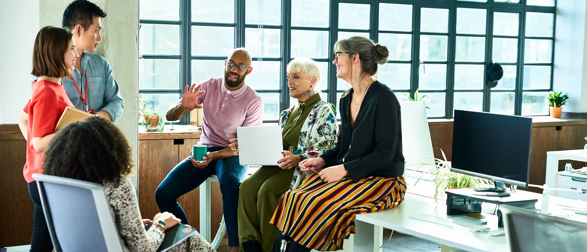 A small group of individuals sitting and standing in a bright, open office, talking together near desks and large windows.