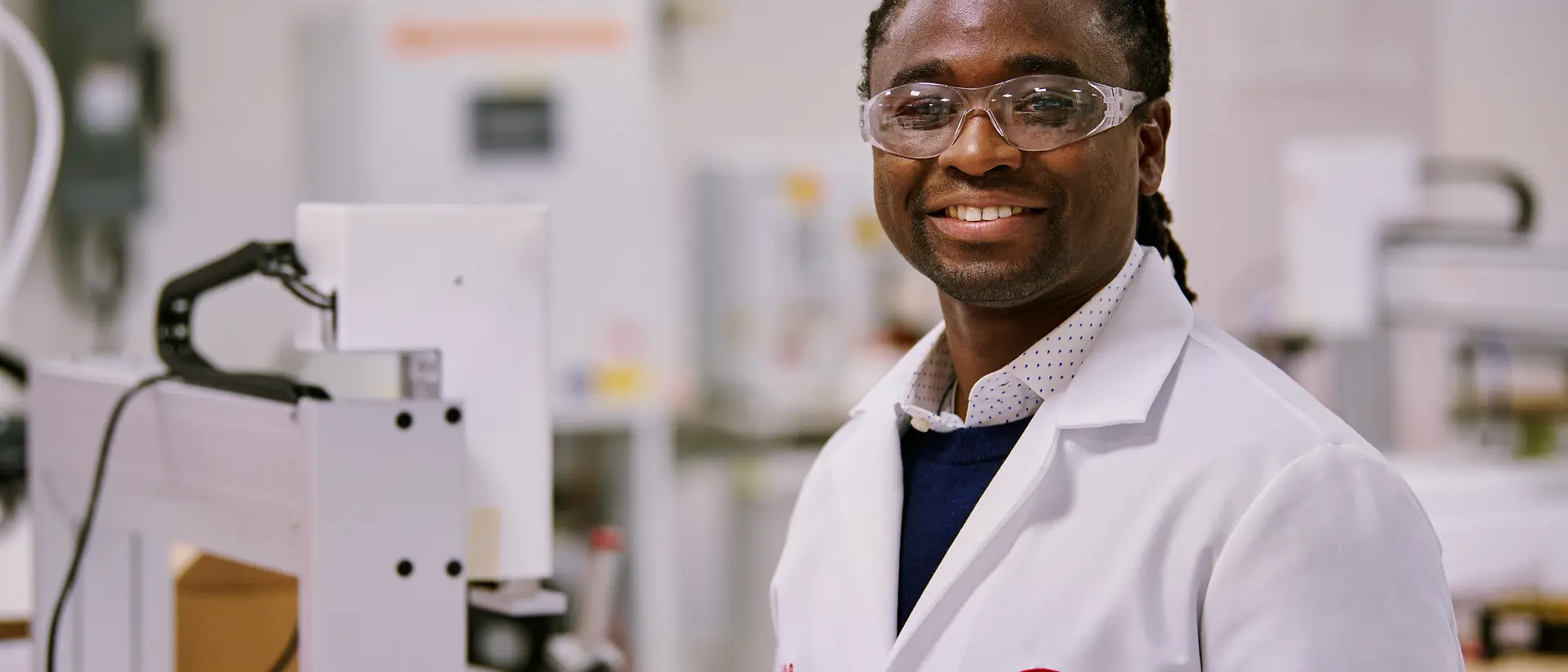 A person in a white Henkel lab coat standing in a laboratory environment surrounded by equipment and machinery.