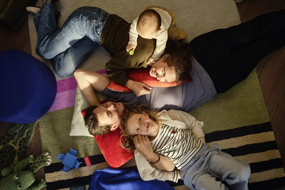 Eine Familie mit Vater, Mutter, Tochter und Baby liegt eng beieinander auf Teppichen und Kissen in einem Wohnraum. Das Baby sitzt auf einem Elternteil, während die anderen Familienmitglieder daneben liegen. Im Umfeld sind Spielzeuge zu sehen.
