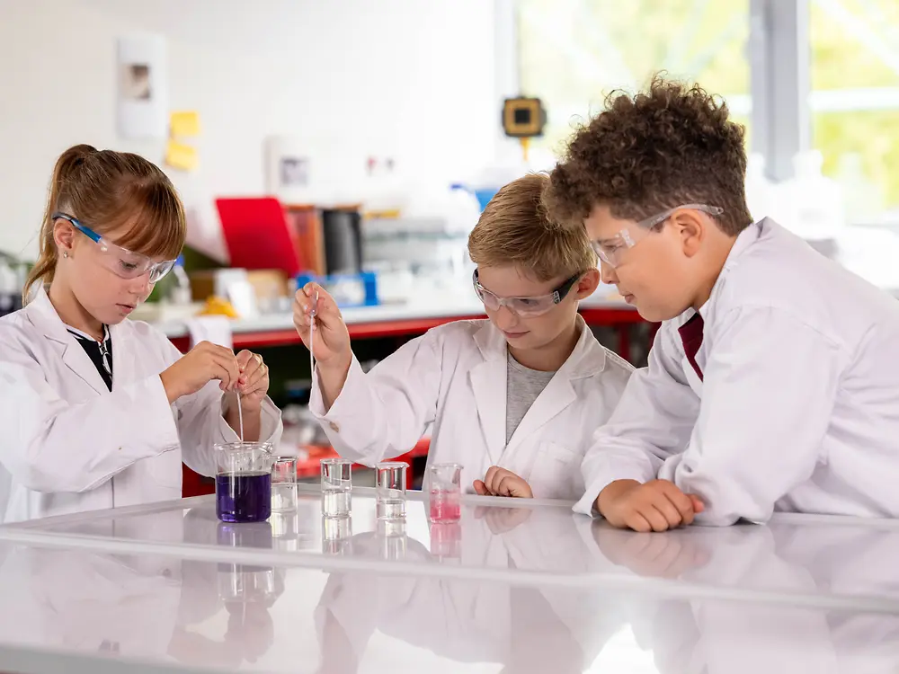 Drei Kinder in Laborkitteln und Schutzbrillen führen gemeinsam ein Experiment mit farbigen Flüssigkeiten durch.